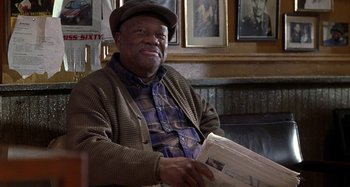Movie still from “Barbershop” (2002), directed by Tim Story – An older man sitting in front of a wall holding a newspaper; Medium shot, Low angle