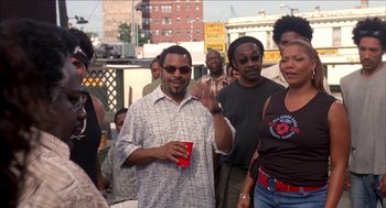 Movie still from “Barbershop 2: Back in Business” (2004), directed by Kevin Rodney Sullivan – A man holding a red cup in front of a group of people; Medium shot, Over the shoulder angle