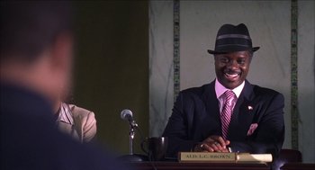 Movie still from “Barbershop 2: Back in Business” (2004), directed by Kevin Rodney Sullivan – A man wearing a suit and hat sitting in front of a microphone; Medium shot, Over the shoulder angle