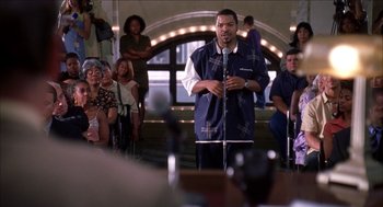Movie still from “Barbershop 2: Back in Business” (2004), directed by Kevin Rodney Sullivan – A man standing at a microphone in front of a group of people; Medium shot, Over the shoulder angle