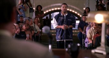 Movie still from “Barbershop 2: Back in Business” (2004), directed by Kevin Rodney Sullivan – A man standing on a stage holding a microphone; Medium shot, Over the shoulder angle