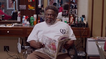 Movie still from “Barbershop: The Next Cut” (2016), directed by Malcolm D. Lee – A man sitting in a barber chair reading a magazine; Medium shot, Over the shoulder angle