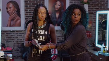 Movie still from “Barbershop: The Next Cut” (2016), directed by Malcolm D. Lee – Two young women standing next to each other holding papers; Medium shot, Over the shoulder angle