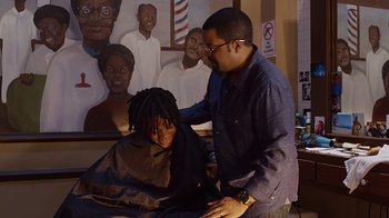 Movie still from “Barbershop: The Next Cut” (2016), directed by Malcolm D. Lee – A man cutting a woman's hair in front of a painting; Medium shot, Over the shoulder angle