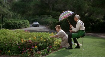Movie still from “Batman: The Movie” (1966), directed by Leslie H. Martinson – An older couple kneeling in the grass while holding an umbrella; Wide shot, High angle