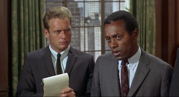 Movie still from “Batman: The Movie” (1966), directed by Leslie H. Martinson – Two men in suits looking at a piece of paper in front of a window; Close Up shot, Over the shoulder angle