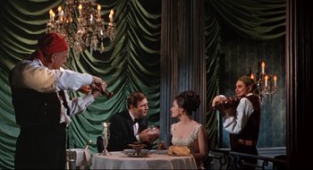Movie still from “Batman: The Movie” (1966), directed by Leslie H. Martinson – A group of people sitting at a table with food; Wide shot, Low angle