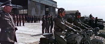 Movie still from “The Battle of Britain” (1969), directed by Guy Hamilton – A group of men standing next to each other on a motorcycle; Wide shot, Low angle