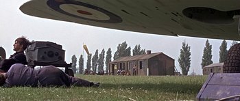 Movie still from “The Battle of Britain” (1969), directed by Guy Hamilton – A group of people sitting in the grass near an airplane; Extreme Wide shot, Low angle