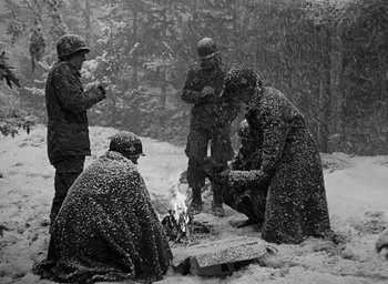 Movie still from “Battleground” (1949), directed by William A. Wellman – A black and white photo of a group of men sitting around a fire in the snow; Wide shot, High angle