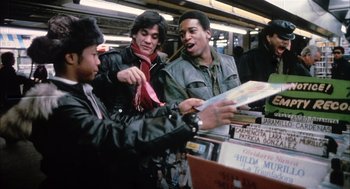 Movie still from “Beat Street” (1984), directed by Stan Lathan – A group of people standing around looking at records on a shelf; Medium shot, High angle
