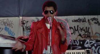Movie still from “Beat Street” (1984), directed by Stan Lathan – A man in a red jacket singing into a microphone; Medium shot, Low angle