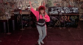 Movie still from “Beat Street” (1984), directed by Stan Lathan – A woman is singing into a microphone in front of a wall covered in graffiti; Wide shot, Low angle