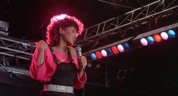 Movie still from “Beat Street” (1984), directed by Stan Lathan – A woman in a pink shirt is holding a microphone; Medium shot, Low angle