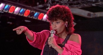 Movie still from “Beat Street” (1984), directed by Stan Lathan – A woman in a pink shirt is holding a microphone; Medium shot, Over the shoulder angle