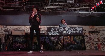 Movie still from “Beat Street” (1984), directed by Stan Lathan – A man standing on a stage holding a microphone; Wide shot, Low angle