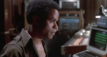 Movie still from “Beat Street” (1984), directed by Stan Lathan – A man sitting in front of a computer in a room; Close Up shot, Over the shoulder angle