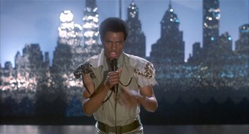 Movie still from “Beat Street” (1984), directed by Stan Lathan – A man with a microphone in front of a city skyline; Medium shot, Low angle