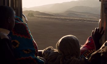 Movie still from “Beau Travail” (1999), directed by Claire Denis – A person sitting in a vehicle on the side of a road; Wide shot, Over the shoulder angle