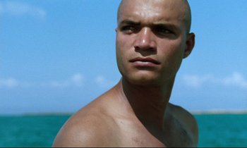 Movie still from “Beau Travail” (1999), directed by Claire Denis – A man with a shaved head is standing in the water; Close Up shot, Low angle