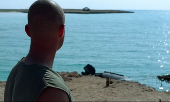 Movie still from “Beau Travail” (1999), directed by Claire Denis – A man standing on the beach looking out at the water; Close Up shot, Over the shoulder angle
