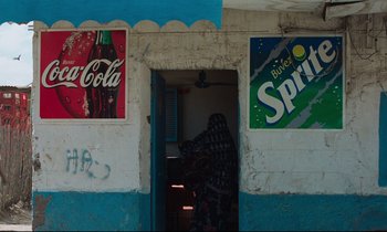 Movie still from “Beau Travail” (1999), directed by Claire Denis – A woman is standing in the doorway of a store; Wide shot, Low angle