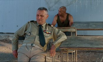 Movie still from “Beau Travail” (1999), directed by Claire Denis – A man in a uniform sitting next to another man in a tank top; Medium shot, Over the shoulder angle