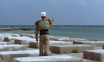 Movie still from “Beau Travail” (1999), directed by Claire Denis – A man in a military uniform is playing a trumpet; Extreme Wide shot, Over the shoulder angle