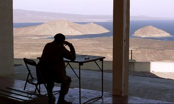 Movie still from “Beau Travail” (1999), directed by Claire Denis – A man sitting at a table in front of an ocean view; Wide shot, High angle
