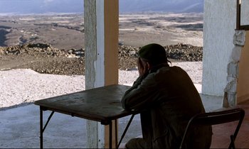 Movie still from “Beau Travail” (1999), directed by Claire Denis – A man sitting at a table with a view of a valley; Wide shot, Over the shoulder angle