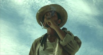 Movie still from “Before Night Falls” (2000), directed by Julian Schnabel – A man with a hat smoking a cigarette in front of a cloudy sky; Medium shot, Low angle