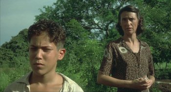 Movie still from “Before Night Falls” (2000), directed by Julian Schnabel – A woman and a young boy standing next to each other in front of trees; Close Up shot, Low angle