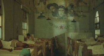 Movie still from “Before Night Falls” (2000), directed by Julian Schnabel – A man sitting on a bed in front of a mural of che guevara; Wide shot, Low angle