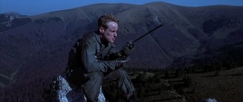 Movie still from “Behind Enemy Lines” (2001), directed by John Moore – A man with a backpack is sitting on a hill holding a rifle; Medium shot, Low angle