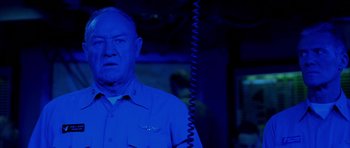 Movie still from “Behind Enemy Lines” (2001), directed by John Moore – An older man in a blue shirt is standing in front of a phone; Close Up shot, Low angle