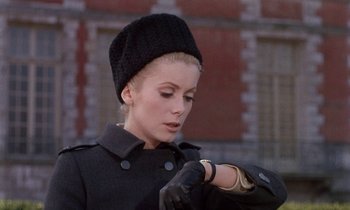 Movie still from “Belle de Jour” (1967), directed by Luis Buñuel – A woman in a black coat and a hat looking at her watch; Close Up shot, Low angle