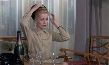 Movie still from “Belle de Jour” (1967), directed by Luis Buñuel – A woman sitting in front of a table with wine glasses on top of her head; Medium shot, Over the shoulder angle