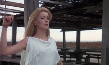 Movie still from “Belle de Jour” (1967), directed by Luis Buñuel – A woman in a white dress standing in front of a wooden structure; Medium shot, Low angle