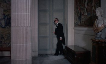 Movie still from “Belle de Jour” (1967), directed by Luis Buñuel – A man in a suit and sunglasses walking through a doorway; Wide shot, Low angle