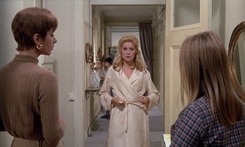 Movie still from “Belle de Jour” (1967), directed by Luis Buñuel – A woman wearing a robe in a hallway with other people; Medium shot, Over the shoulder angle