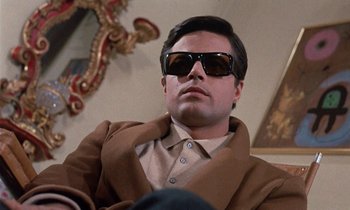 Movie still from “Belle de Jour” (1967), directed by Luis Buñuel – A man wearing sunglasses sitting in a room; Close Up shot, Low angle