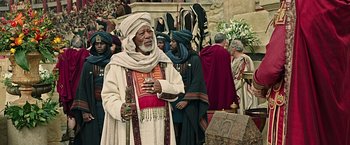 Movie still from “Ben-Hur” (2016), directed by Timur Bekmambetov – An older man in a costume standing in front of a group of people; Medium shot, Over the shoulder angle