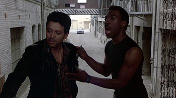 Movie still from “Beverly Hills Cop” (1984), directed by Martin Brest – A man is combing another man's hair in the street; Medium shot, Over the shoulder angle