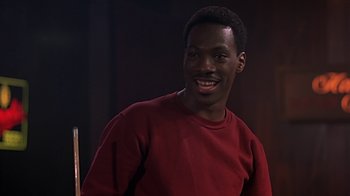 Movie still from “Beverly Hills Cop” (1984), directed by Martin Brest – A man in a red shirt smiling for the camera; Close Up shot, Over the shoulder angle