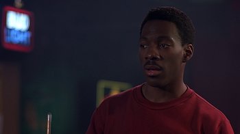 Movie still from “Beverly Hills Cop” (1984), directed by Martin Brest – A man in a red t - shirt is looking at the camera; Close Up shot, Over the shoulder angle