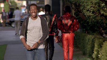 Movie still from “Beverly Hills Cop” (1984), directed by Martin Brest – A man smiles while walking down the street; Medium shot, Over the shoulder angle