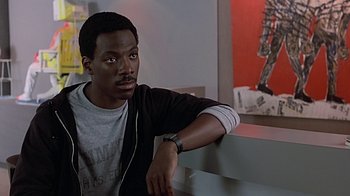 Movie still from “Beverly Hills Cop” (1984), directed by Martin Brest – A man leaning against a wall in a room; Close Up shot, Over the shoulder angle