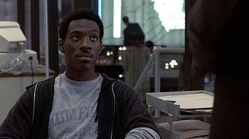Movie still from “Beverly Hills Cop” (1984), directed by Martin Brest – A man sitting at a table in front of a projection screen; Close Up shot, Over the shoulder angle