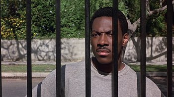 Movie still from “Beverly Hills Cop” (1984), directed by Martin Brest – A man sitting behind a metal fence looking at the camera; Close Up shot, Low angle