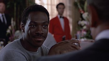 Movie still from “Beverly Hills Cop” (1984), directed by Martin Brest – A person sitting at a table in a room; Close Up shot, Over the shoulder angle